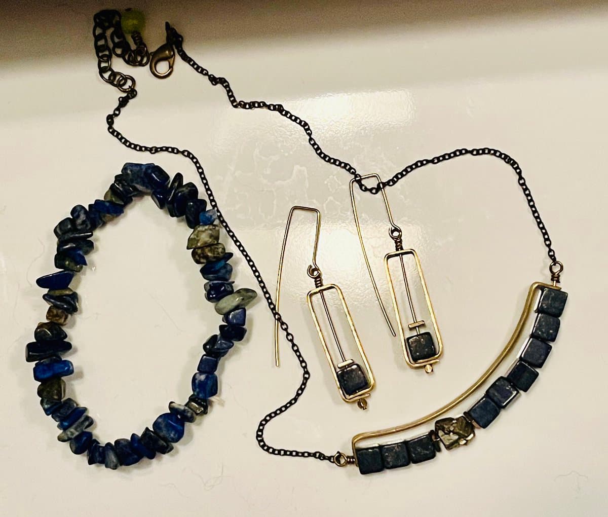 A flat-lay photograph of a lapis lazuli jewelry set, including a beaded bracelet, a beaded necklace with a gold-tone clasp, and a pair of rectangular drop earrings, all arranged on a white surface.