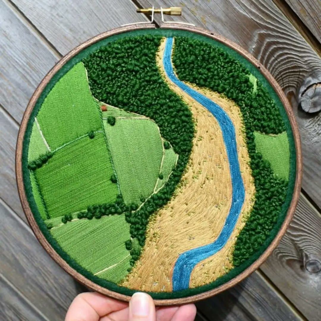 A close-up photograph of a circular embroidery hoop held by a hand, displaying a stitched aerial map of a landscape with green fields, a forest, and a blue river, set against a weathered wooden background.