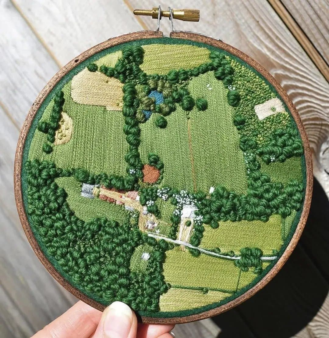 A close-up photograph of a hand holding a wooden embroidery hoop containing a stitched, aerial map of a rural landscape. The piece features textured greenery, fields, a pond, and small buildings, set against a background of light wooden planks.