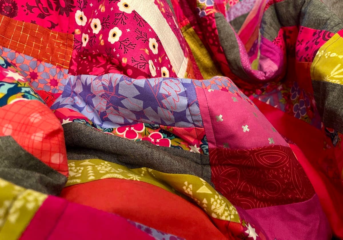 A close-up photograph of a colorful patchwork quilt featuring various fabric patterns in shades of pink, red, purple, and green. The image highlights the texture and stitching of the different fabric squares sewn together.