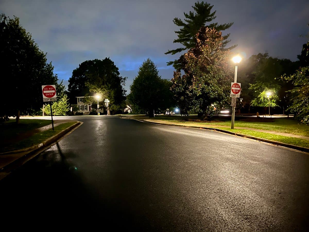 A nighttime photograph of a wet, empty residential street with streetlights and trees. Two red 'DO NOT ENTER' signs are visible on either side of the road ahead.