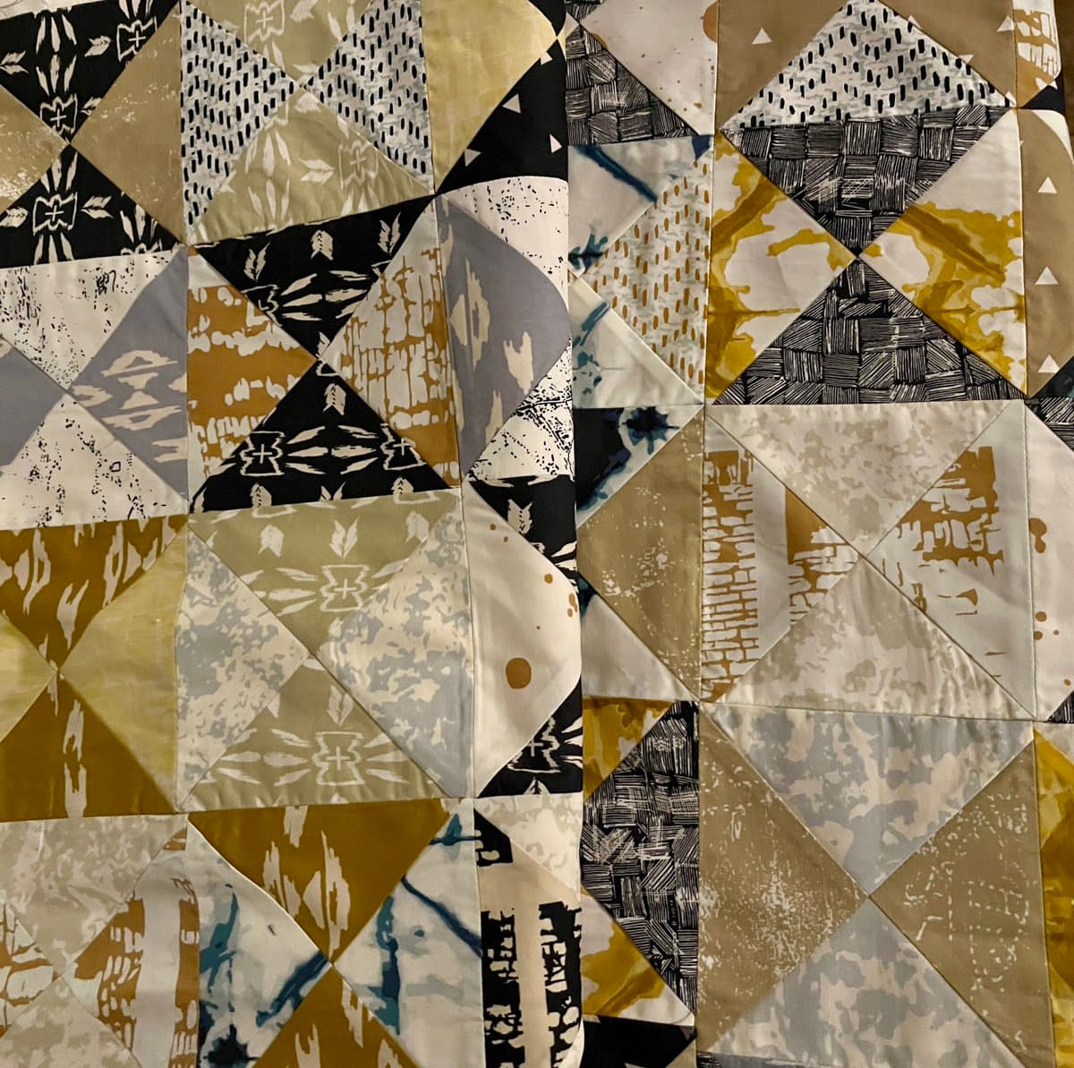 A close-up photograph of a patchwork quilt featuring a geometric pattern of triangles and squares in a color palette of black, white, gold, and gray. The fabrics display various prints, including floral, abstract, and textured designs, stitched together to form a complex, multi-faceted surface.