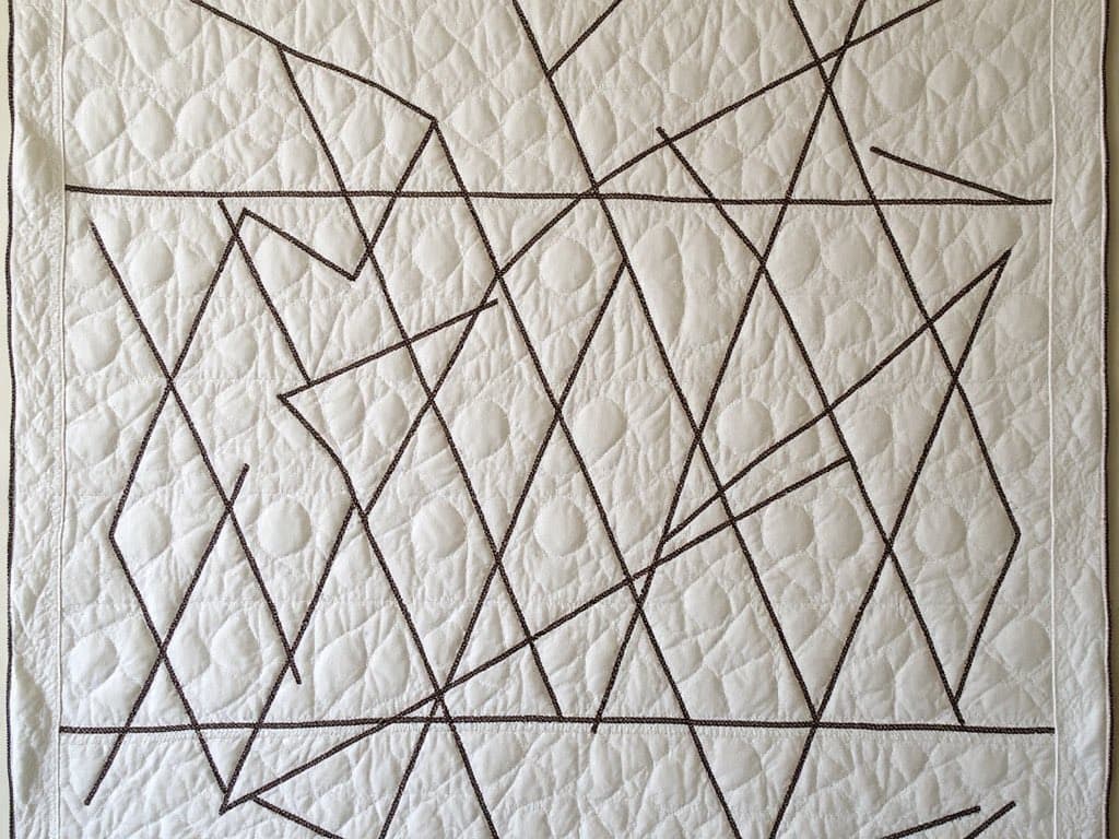 A close-up view of a white quilt featuring a geometric pattern of intersecting dark brown stitched lines. The background fabric has a subtle, repeating circular quilting texture.