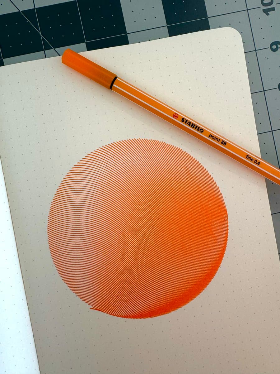 A photograph of a dotted-grid notebook page featuring a large, orange circle drawn with fine, parallel lines, with an orange Stabilo Point 88 pen resting on the page above the drawing.