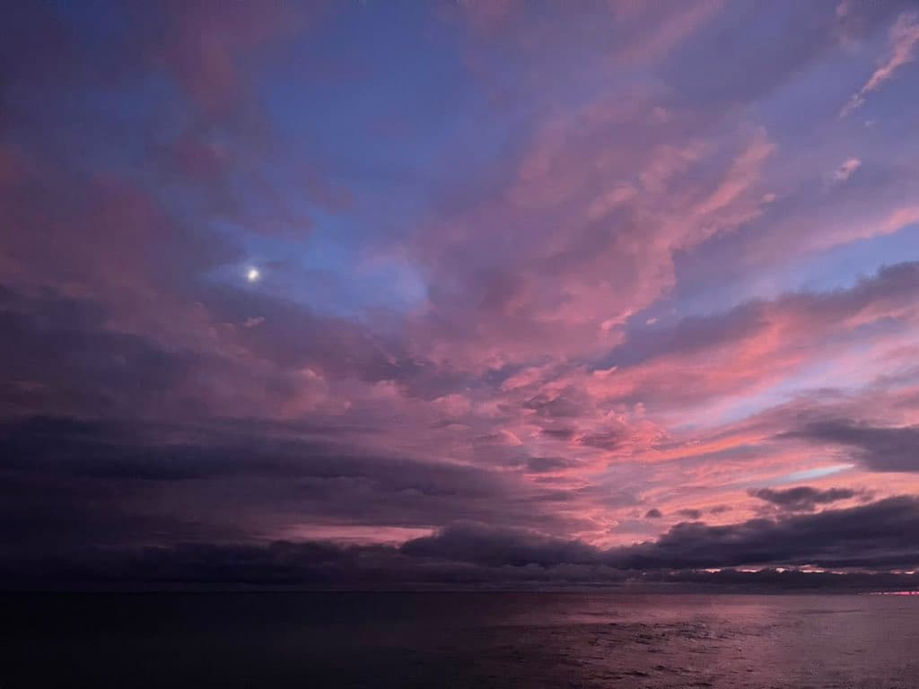 A scenic photograph of a sunset or sunrise over a body of water, featuring a sky filled with clouds illuminated in shades of pink, purple, and blue, with a small bright light visible in the upper left.