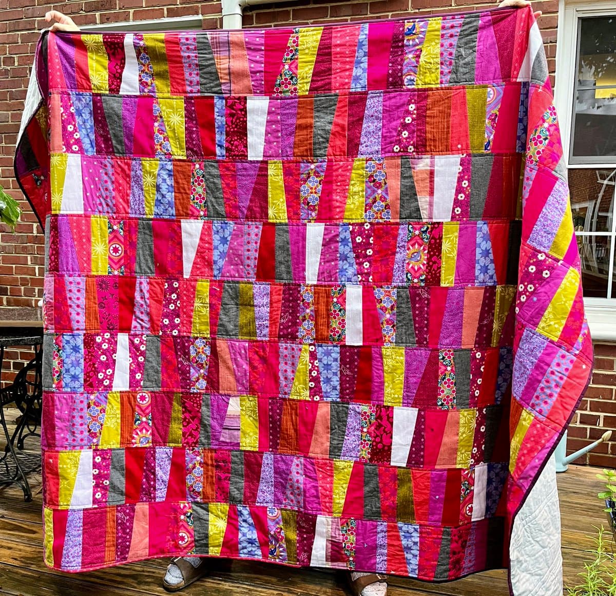 A person holds up a large, colorful patchwork quilt featuring a geometric pattern of vertical strips in shades of pink, red, purple, yellow, and blue. The quilt is displayed outdoors on a wooden deck in front of a brick wall.