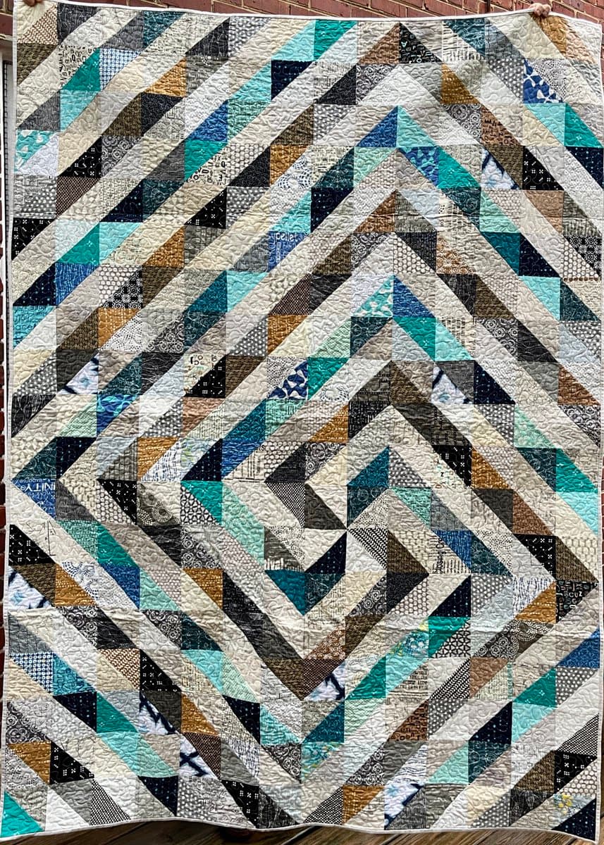A large, finished patchwork quilt hangs against a brick wall, featuring a complex geometric pattern of chevrons and triangles in shades of teal, blue, brown, and cream. The quilt is made from various patterned fabrics and displays visible quilting stitches across its surface.