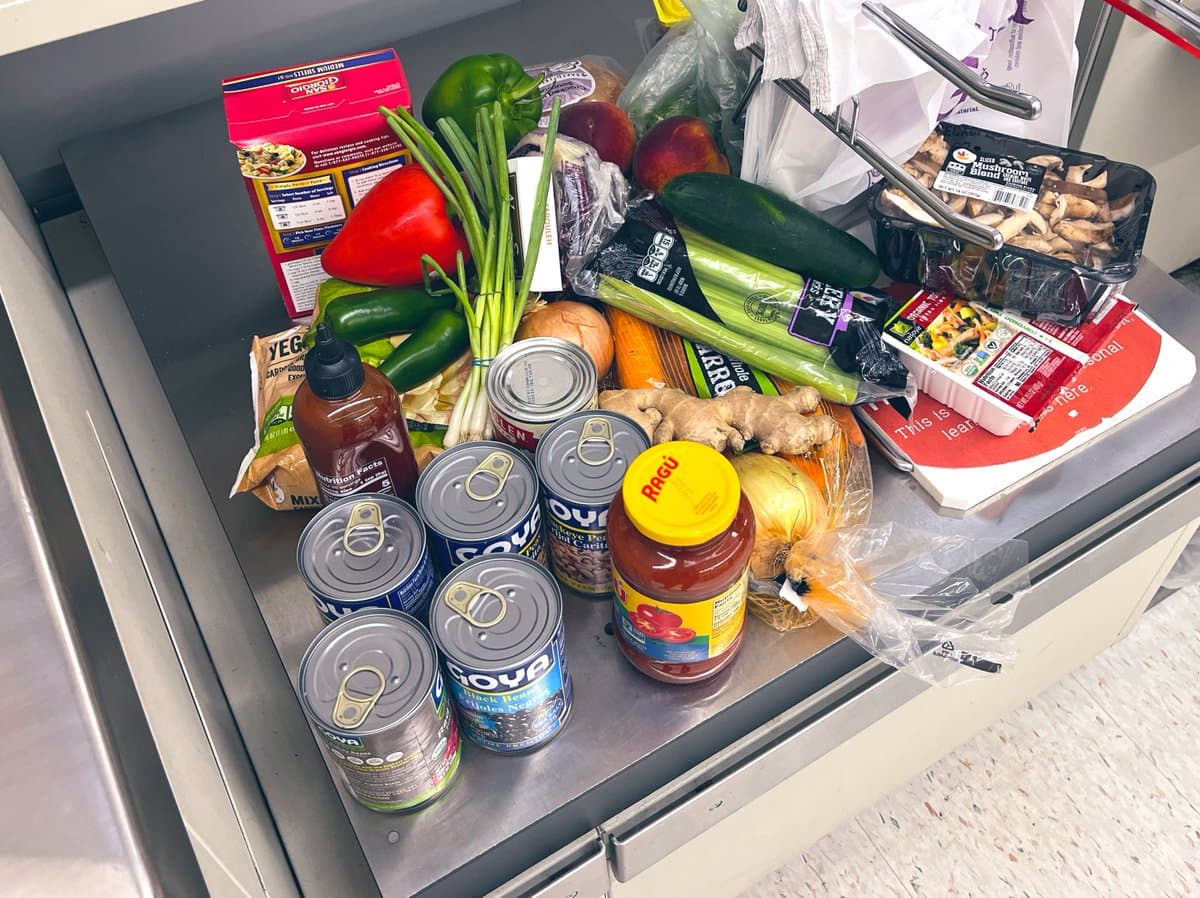 A top-down view of a stainless steel counter filled with a variety of fresh produce and canned goods, including bell peppers, green onions, celery, mushrooms, and cans of Goya beans.