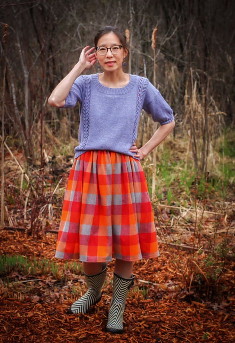 A woman stands outdoors in a wooded area, wearing a purple knit sweater, a bright orange and red plaid skirt, and black-and-white patterned rain boots. She is posing with one hand on her hip and the other near her head.