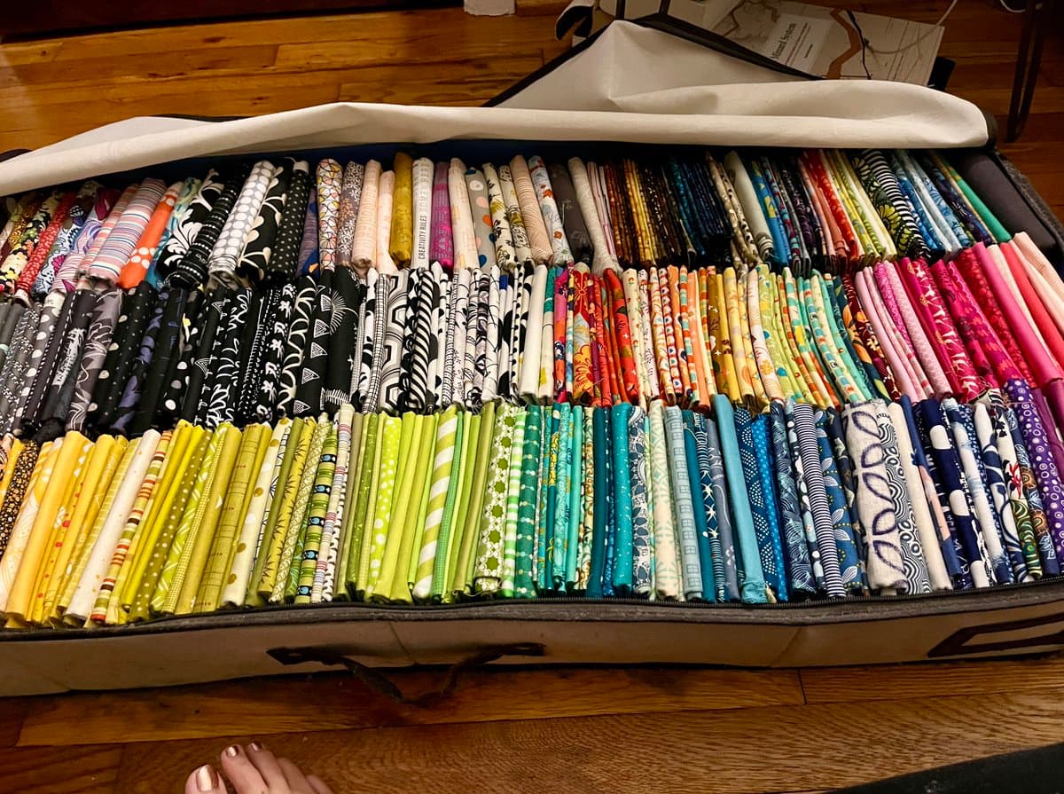 A photo of an open suitcase filled with numerous bolts of fabric, neatly organized by color and pattern. The collection features a wide variety of prints, including floral, geometric, and abstract designs, arranged in rows within the luggage.