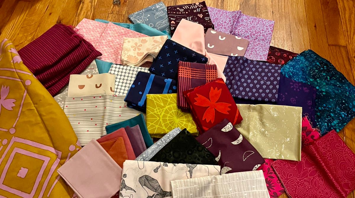 A collection of folded fabric pieces in various colors and patterns is arranged on a wooden floor. The fabrics feature designs such as florals, geometric shapes, and abstract prints, suggesting they are materials for sewing or crafting.