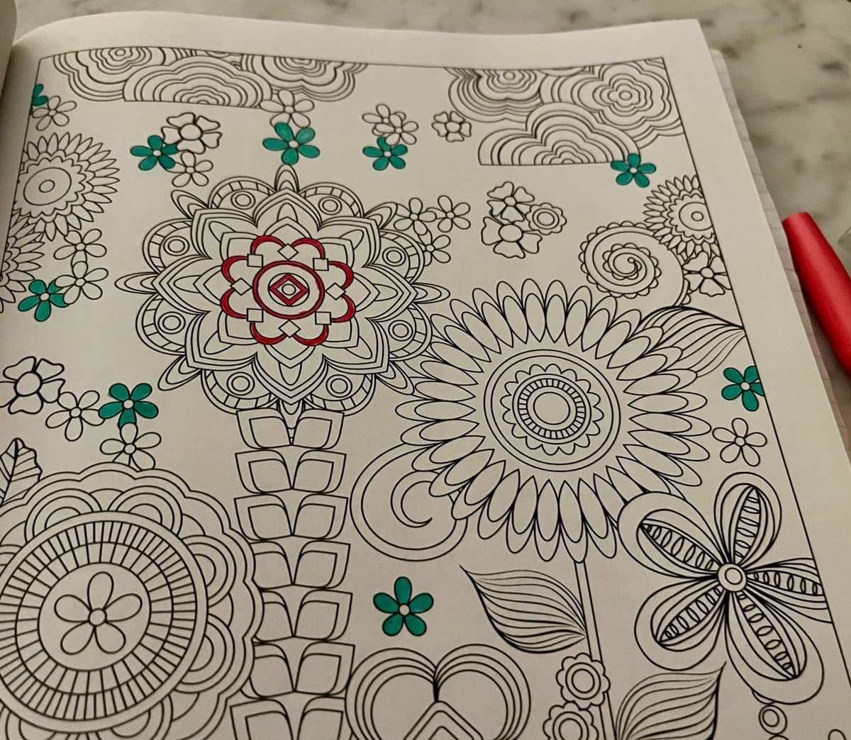 A photograph of an open coloring book page featuring intricate black-and-white line art of flowers and mandalas, with some sections partially colored in red and green. A red coloring pencil rests on the right edge of the page.