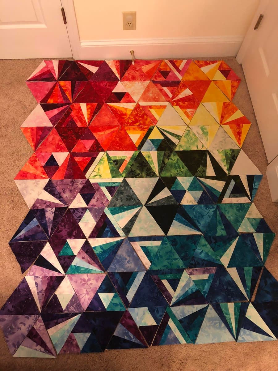 A large, colorful patchwork quilt laid out on a carpeted floor, featuring a geometric pattern of triangles in a rainbow gradient from reds and oranges to purples and blues.
