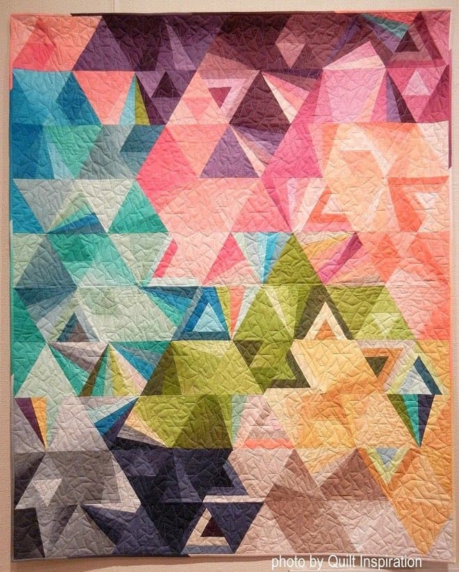 A colorful geometric quilt featuring a complex pattern of triangles in shades of pink, blue, green, and purple, with a credit in the bottom right corner.