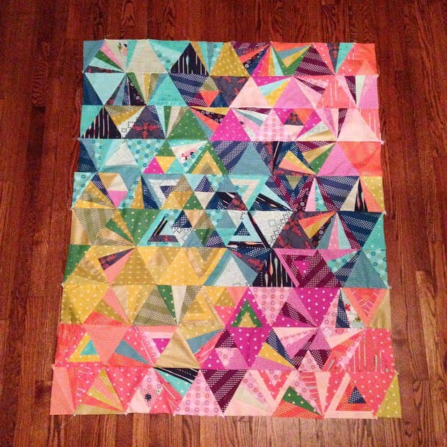 A colorful, geometric quilt top laid flat on a wooden floor, featuring a patchwork of triangular fabric pieces in various patterns and bright colors. The design creates a vibrant, mosaic-like appearance with sections of pink, blue, green, and yellow.