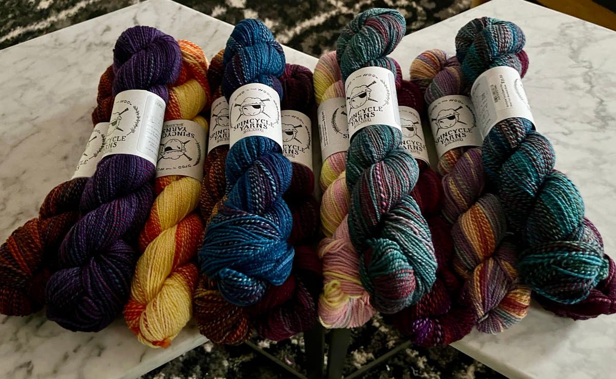 A collection of colorful skeins of yarn from Spincycle Yarns arranged on a marble surface. The skeins feature various colorways, including purple, yellow, blue, and green, with white labels displaying the brand name and a sheep logo.