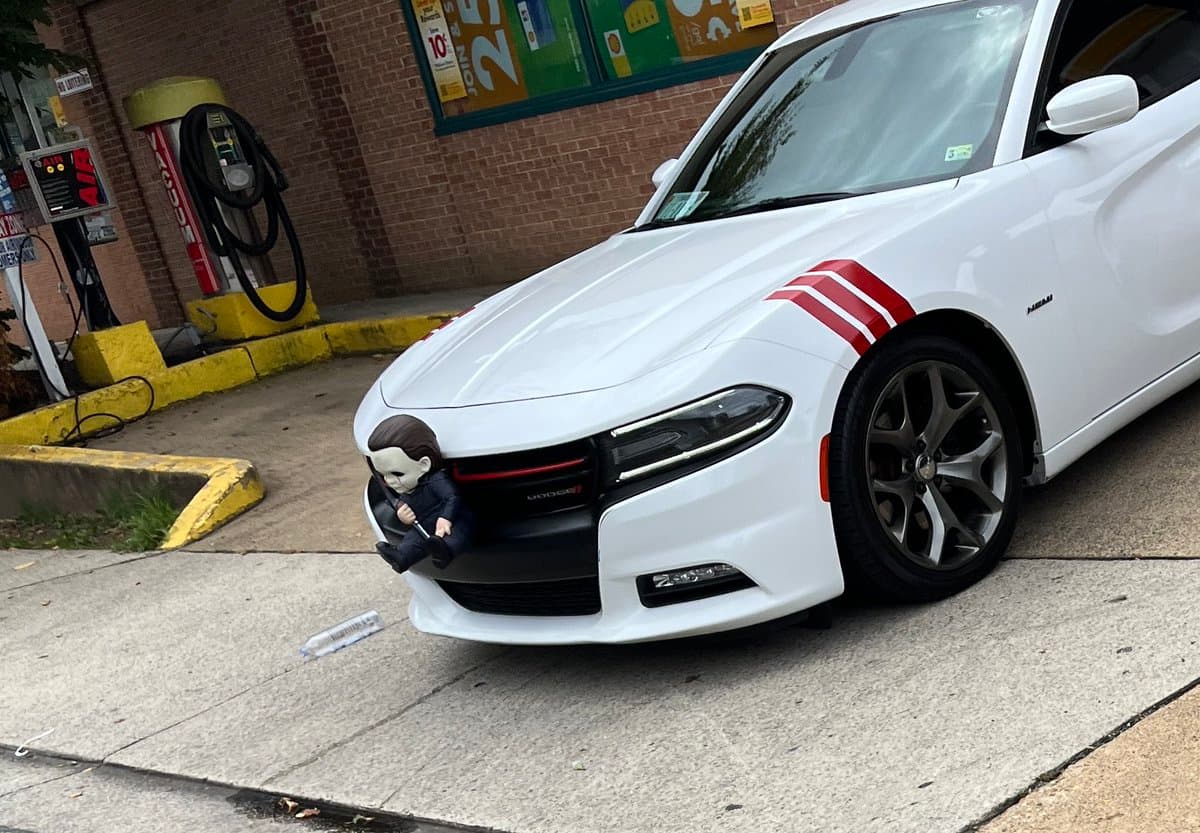 A white Dodge Charger with red racing stripes is parked at a gas station, featuring a Michael Myers action figure sitting in the front grille.