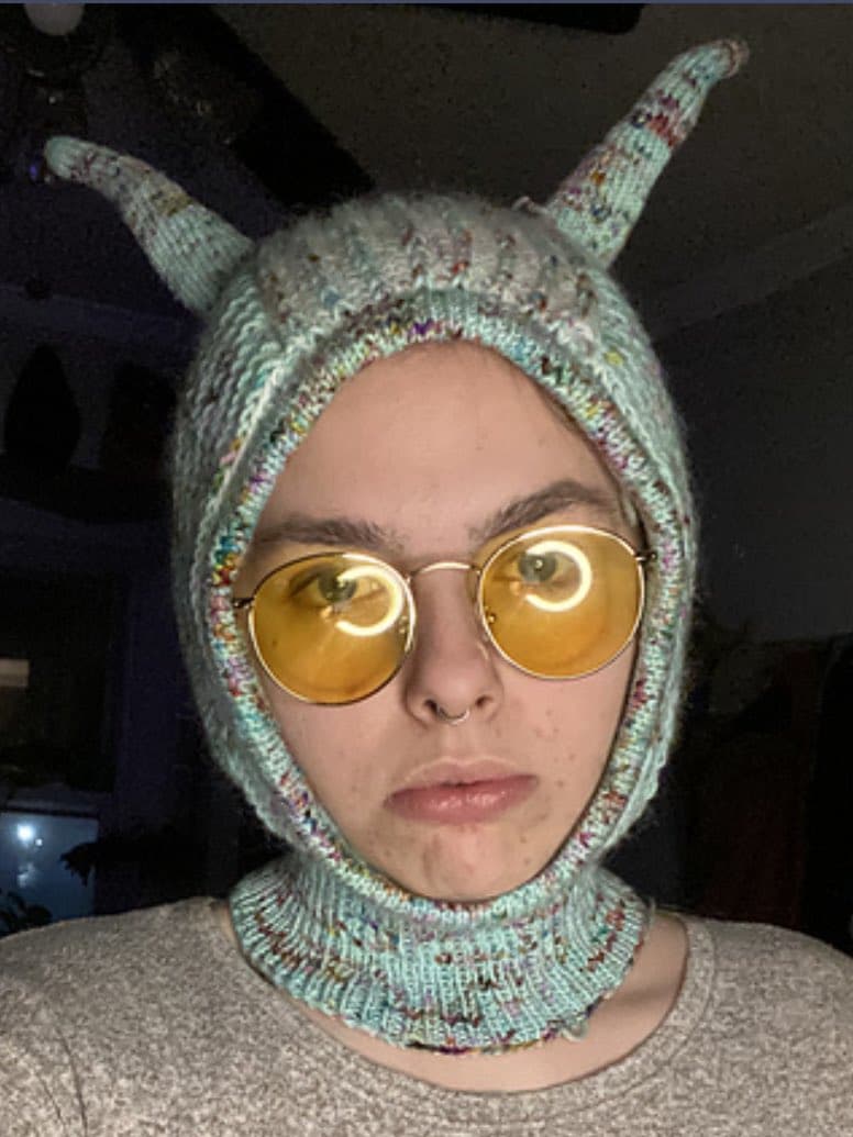 A selfie of a person wearing a light blue, knitted hood with two pointed, ear-like protrusions and round, yellow-tinted glasses. The person has a neutral expression and is wearing a gray shirt.