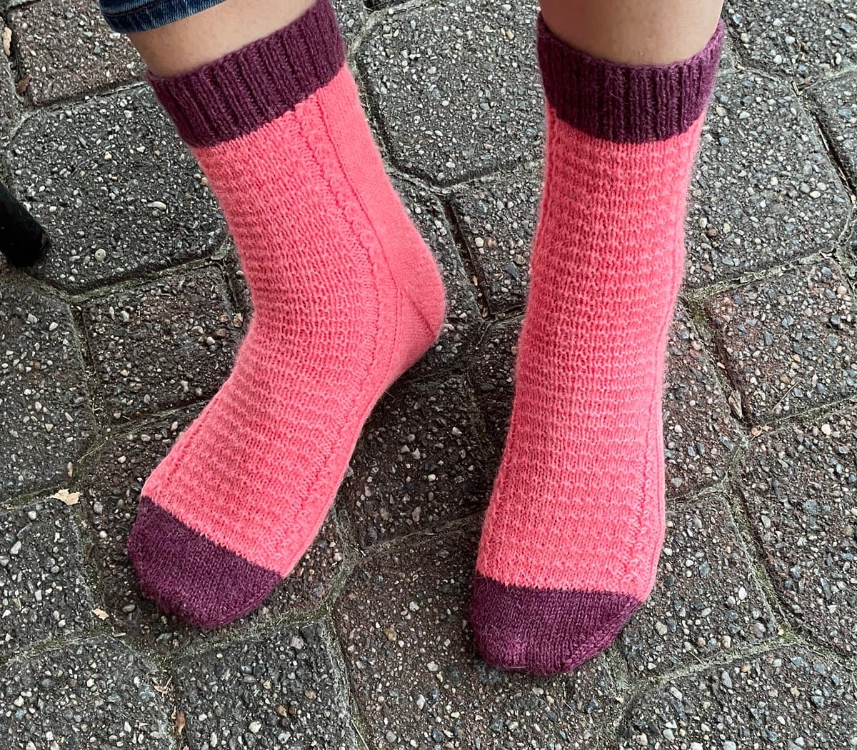 A close-up, top-down photograph of a person's feet wearing a pair of hand-knitted socks. The socks are primarily pink with dark purple cuffs and toe caps, and are shown standing on a paved stone surface.