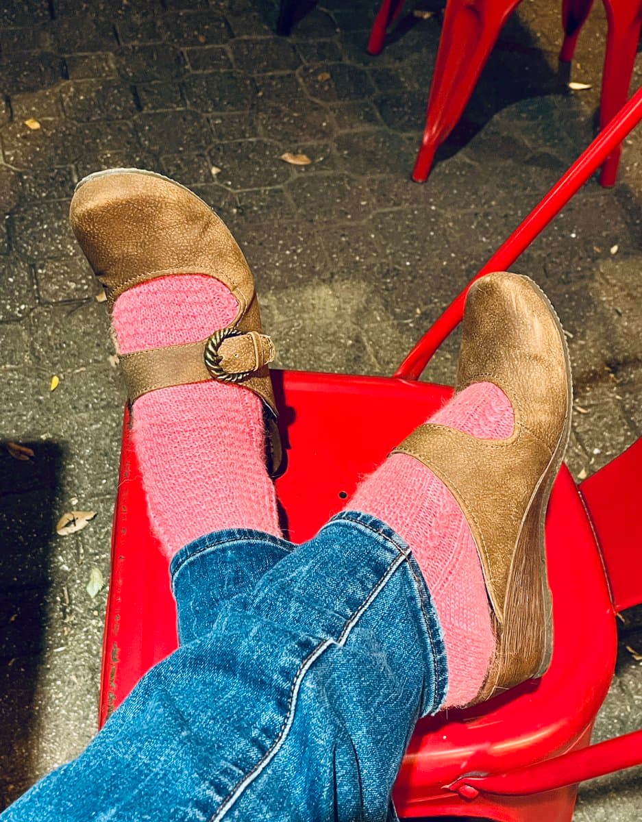 A first-person view of a person's feet wearing pink knitted socks and tan suede clog-style shoes, resting on a red metal chair outdoors.