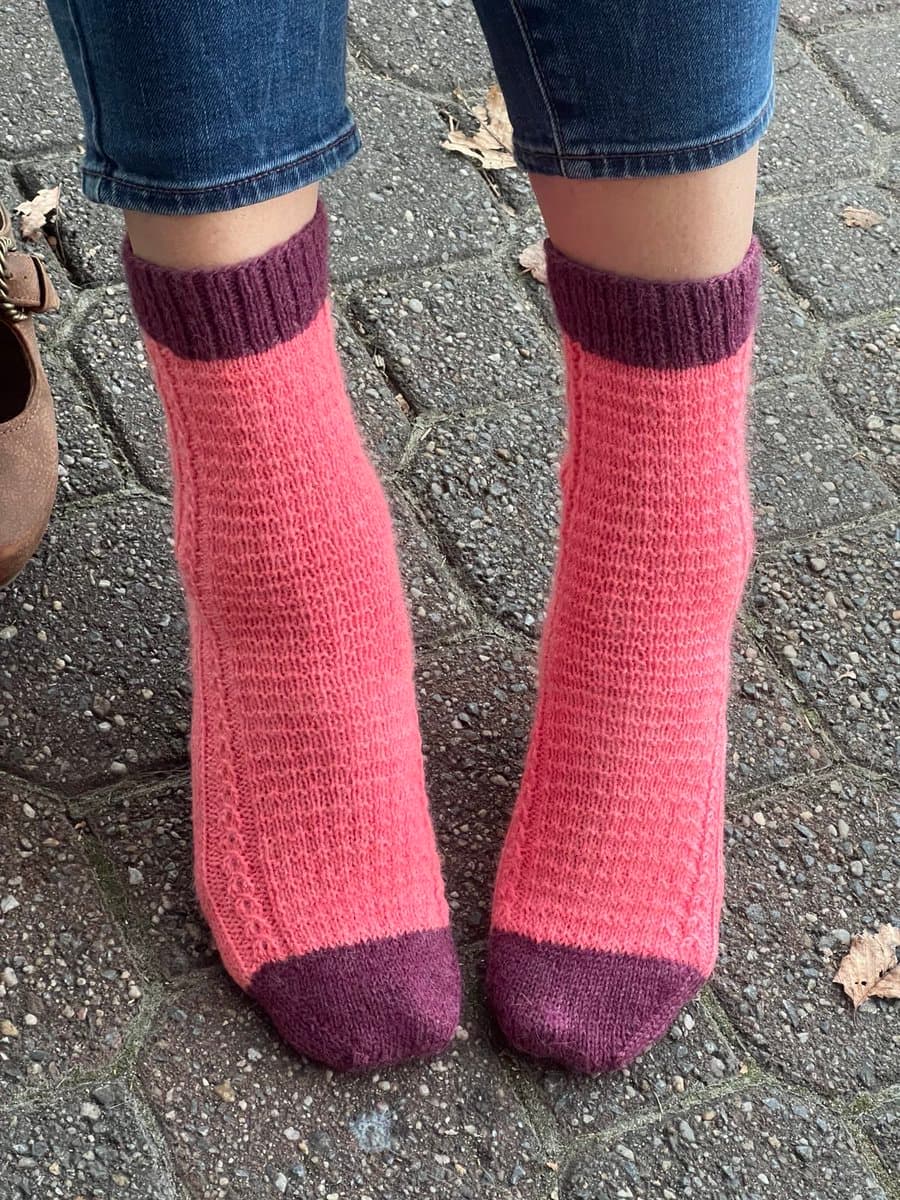 A close-up, low-angle photograph of a person's lower legs and feet standing on a paved surface. The person is wearing blue jeans and a pair of hand-knitted socks that are primarily pink with dark purple cuffs and toe caps.