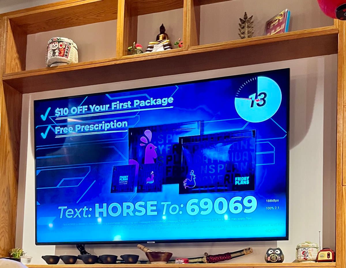 A television screen displays a blue advertisement for a mobile phone plan, featuring a purple horse mascot and a clock showing 13. The TV is mounted on a wall beneath wooden shelves holding various decorative items, including a Buddha statue and a Maneki-neko figurine.