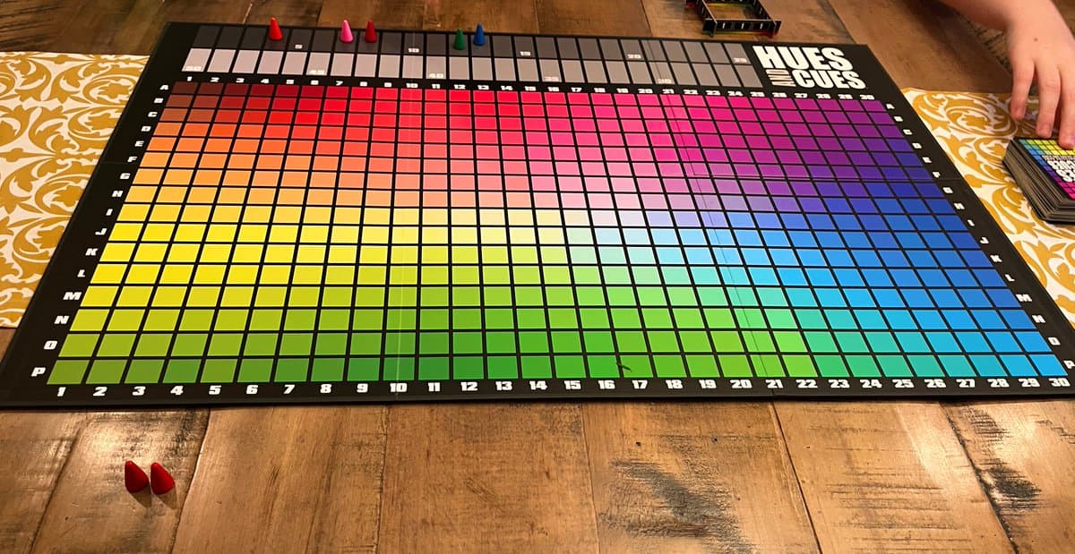A photograph of the board game 'Hues and Cues' laid out on a wooden table, featuring a large grid of colored squares arranged in a rainbow spectrum. Small colored pegs are placed on the top row of the board, and a person's hand is visible on the right side.
