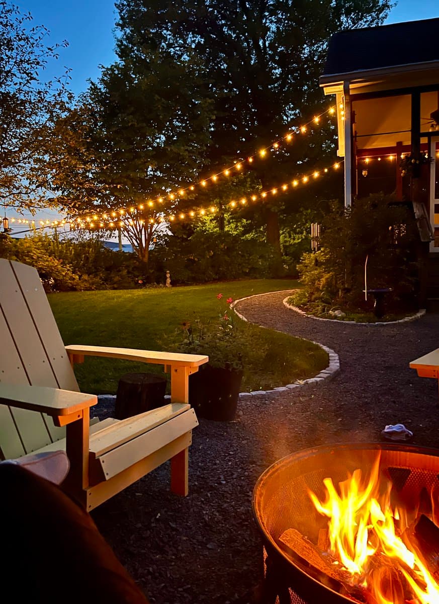 A backyard scene at dusk featuring a lit fire pit in the foreground, a wooden Adirondack chair, and a string of warm lights strung across the yard. A gravel path winds through the green lawn toward a house with a lit porch.