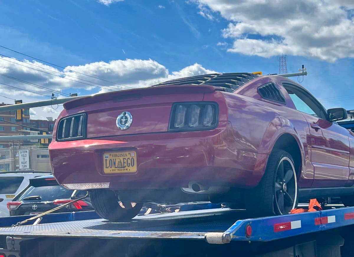 A purple Ford Mustang is being loaded onto a blue flatbed tow truck on a sunny day. The car has a Virginia license plate that reads "LOW AEGO" with the slogan "DON'T TREAD ON ME." In the background, there are other vehicles, buildings, and a street sign that says "U-TURN YIELD TO RIGHT TURN."