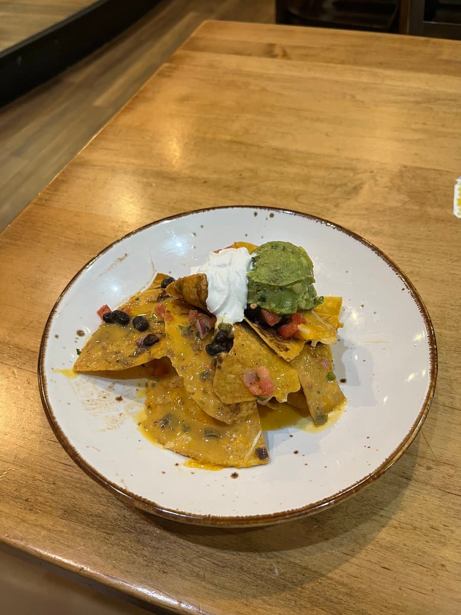A small portion of nachos on a white plate, topped with a dollop of sour cream and a scoop of guacamole, sitting on a wooden table.