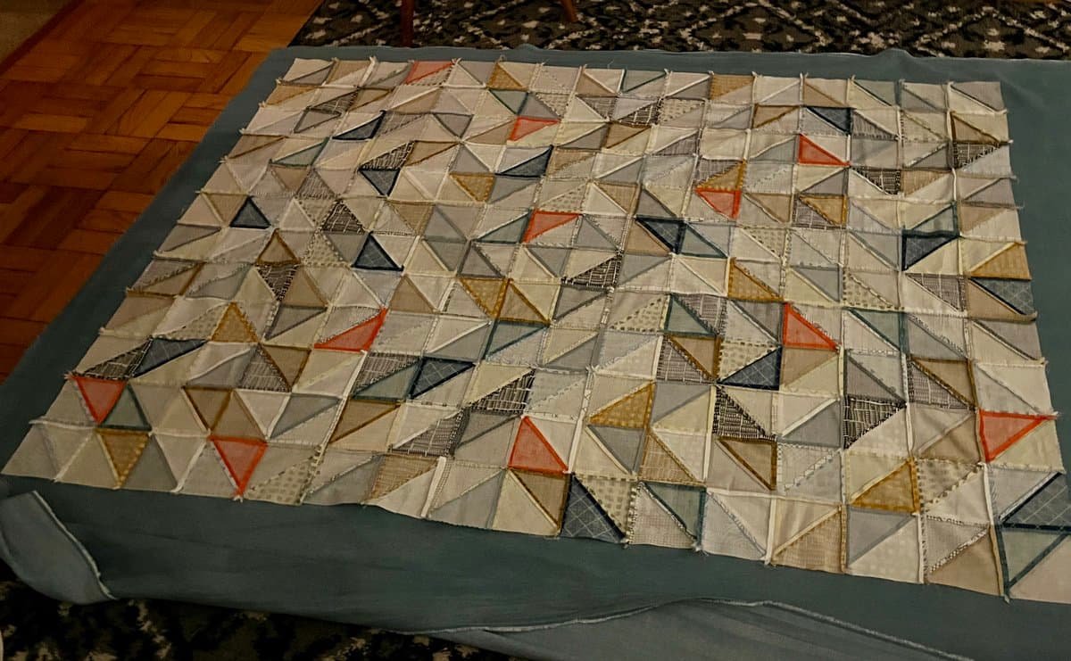 A photograph of a quilt in progress laid out on a bed, featuring a geometric pattern of triangles in shades of white, grey, blue, and orange.
