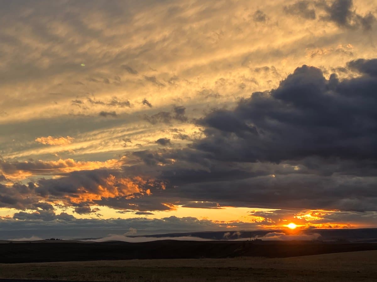 A scenic photograph of a sunset over a rolling landscape, featuring a dramatic sky with dark clouds and golden light. The sun is setting on the horizon, casting a warm glow across the clouds and the distant hills.