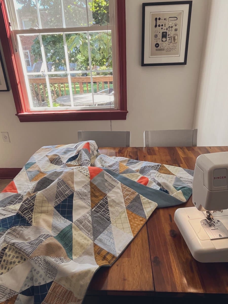A colorful patchwork quilt is spread out on a wooden table next to a white Singer sewing machine, with a window and framed art in the background.