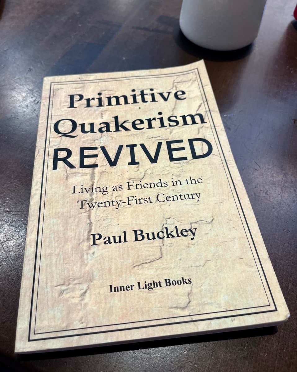 A paperback book titled 'Primitive Quakerism Revived' by Paul Buckley, published by Inner Light Books, is lying on a dark wooden table. The book cover has a textured, light-colored background with black text and a thin border.