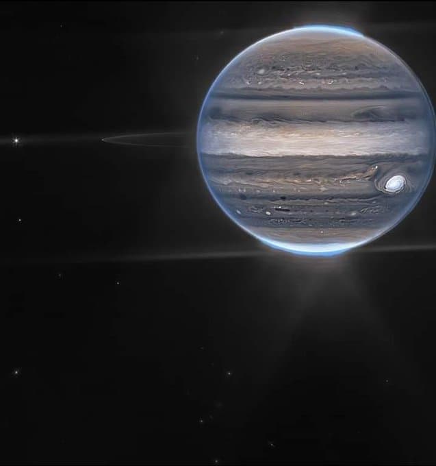 A photograph of the planet Jupiter set against the blackness of space, showing its characteristic banded cloud patterns and the Great Red Spot. The image features a bright blue atmospheric glow along the planet's limb and a horizontal lens flare streaking across the frame.