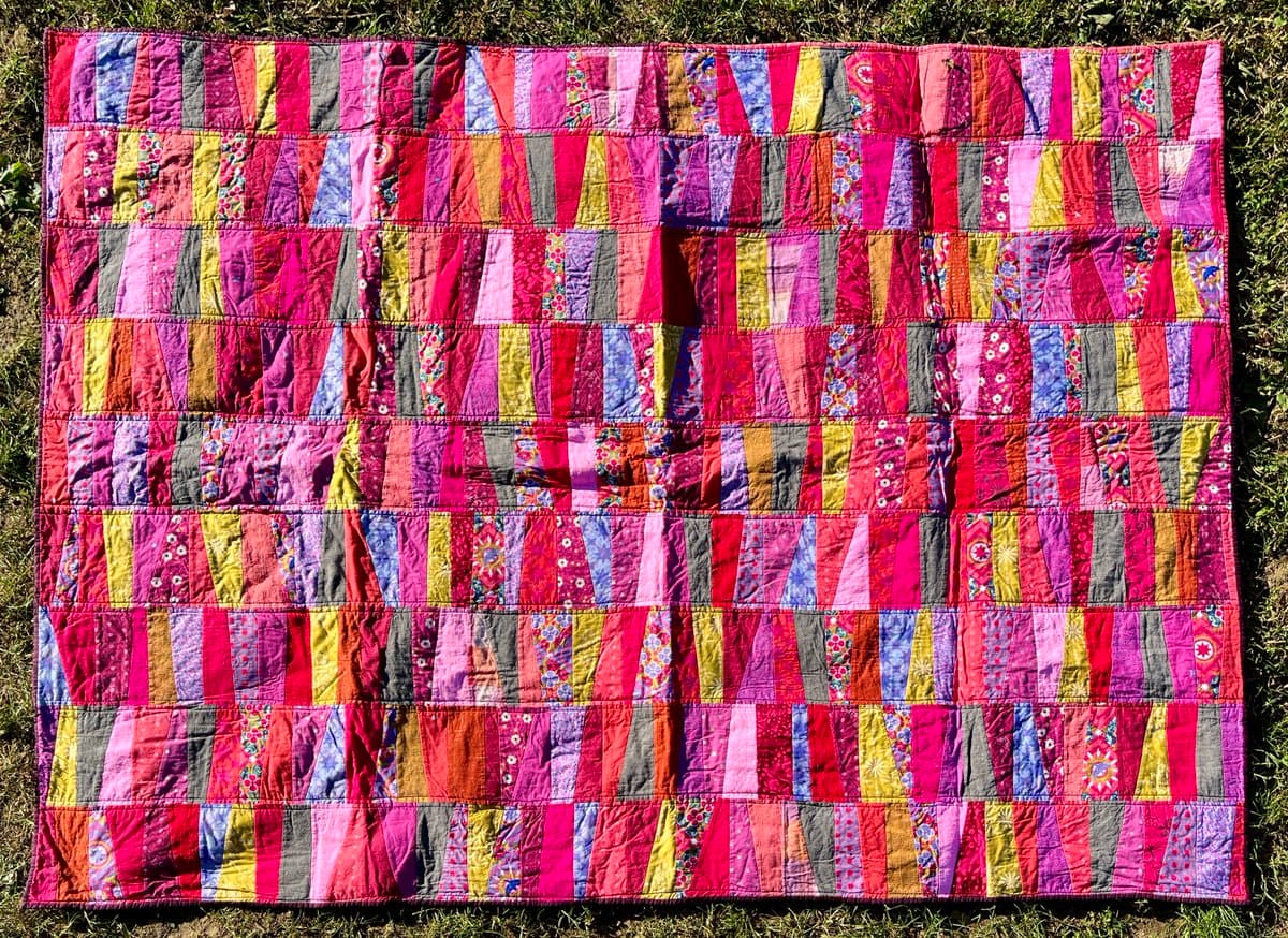A colorful patchwork quilt featuring a grid of vertical stripes in shades of pink, red, purple, and yellow, spread out on a grassy surface.