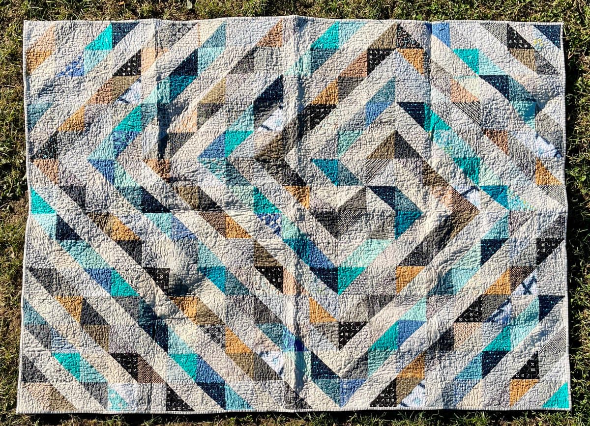 A finished quilt with a geometric pattern of diagonal stripes in shades of blue, teal, brown, and black is laid out on a grassy surface. The quilt features a central diamond design and is displayed outdoors in natural light.