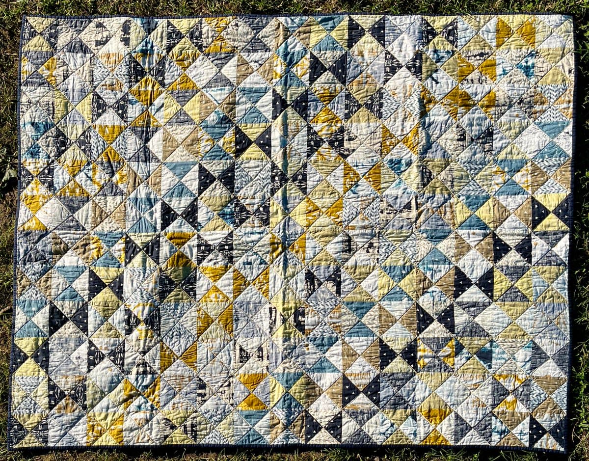 A top-down view of a finished patchwork quilt laid out on a grassy surface. The quilt features a geometric pattern of diamond-shaped blocks in shades of blue, yellow, black, and white, with visible quilting stitches across the surface.