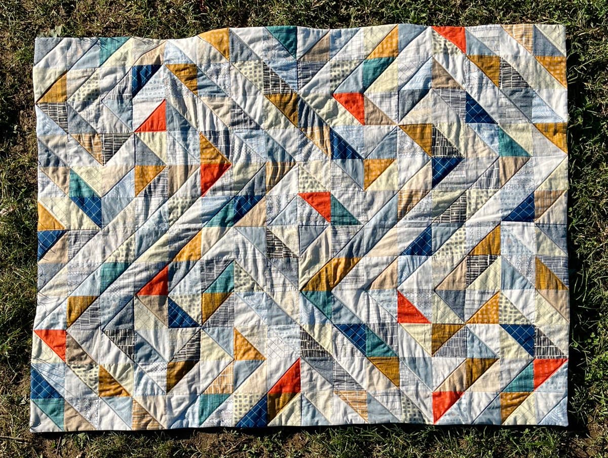 A top-down view of a finished patchwork quilt laid out on a grassy surface. The quilt features a geometric pattern composed of triangles in shades of blue, yellow, orange, and white.