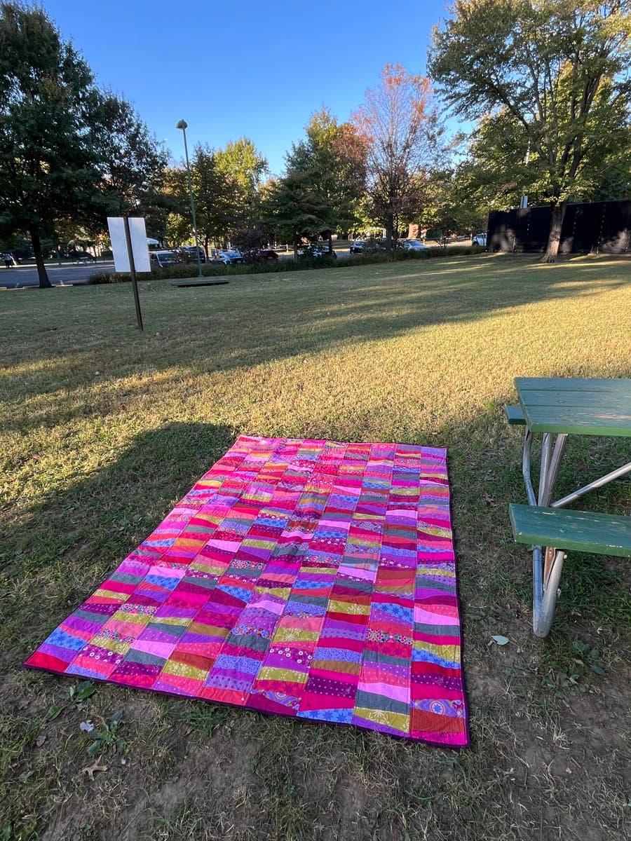 A colorful, patchwork quilt is spread out on a grassy lawn next to a green picnic table, with trees and a parking lot visible in the background under a clear blue sky.