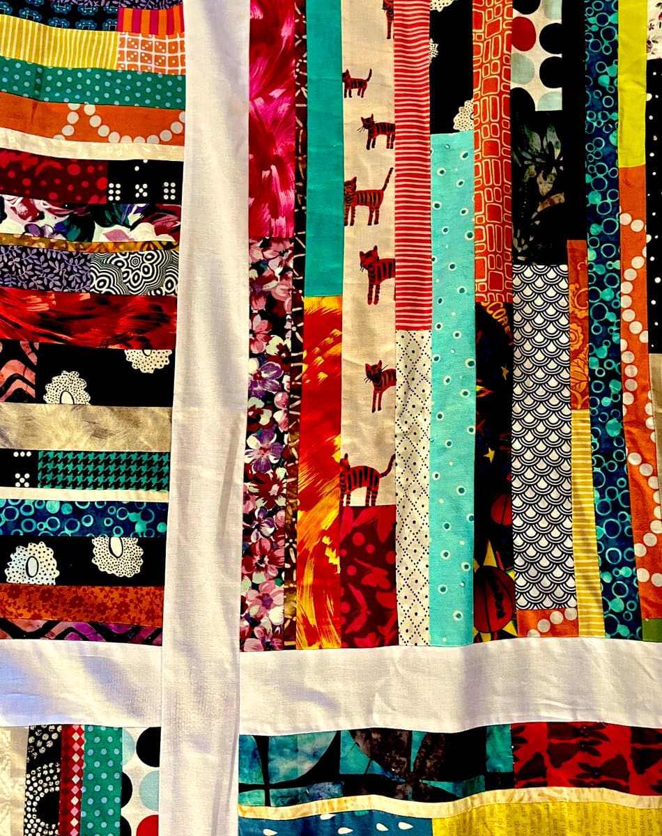 A close-up photograph of a colorful patchwork quilt featuring vertical strips of various patterned fabrics, including a prominent strip with a repeating cat design. The quilt is divided into sections by wide, plain white sashing strips.