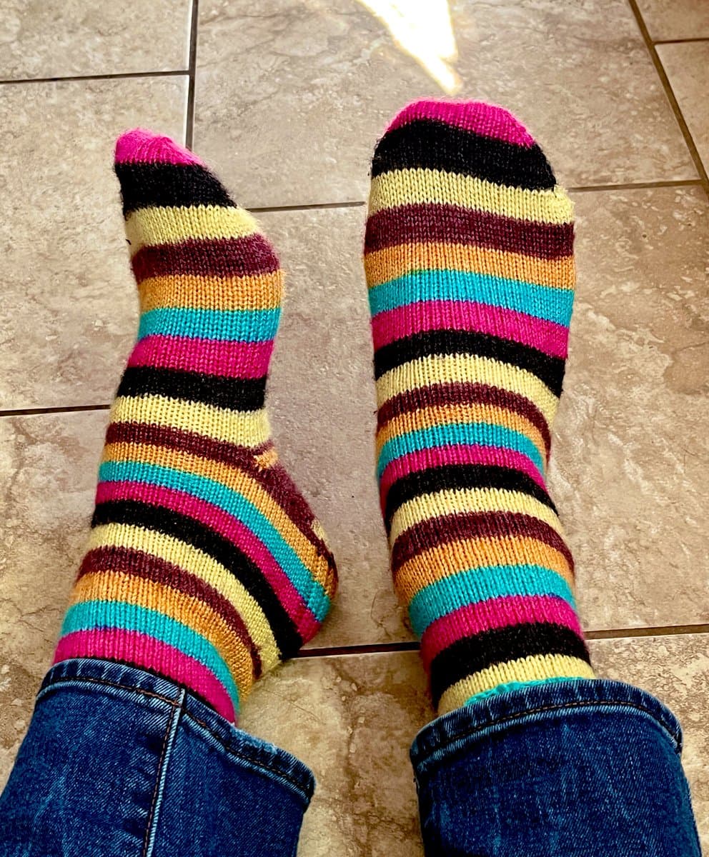 A first-person view of a person's feet wearing colorful, striped hand-knitted socks while standing on a tiled floor. The socks feature horizontal bands of pink, black, yellow, orange, maroon, and teal, and are paired with blue jeans.