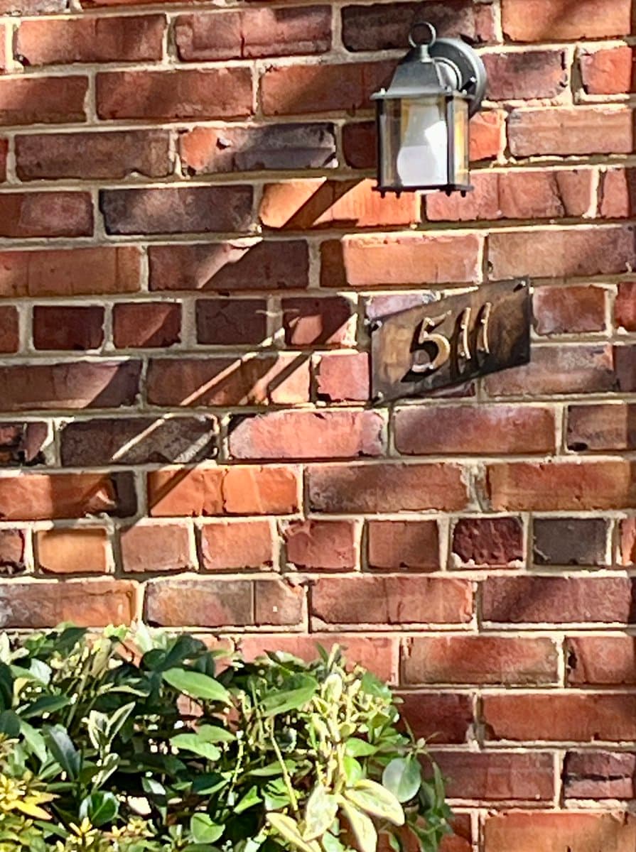 A close-up photograph of a red brick wall featuring a small, dark metal plaque with the house number '511' in low-contrast copper lettering, mounted below a wall-mounted lantern light fixture. Green foliage is visible at the bottom of the frame.