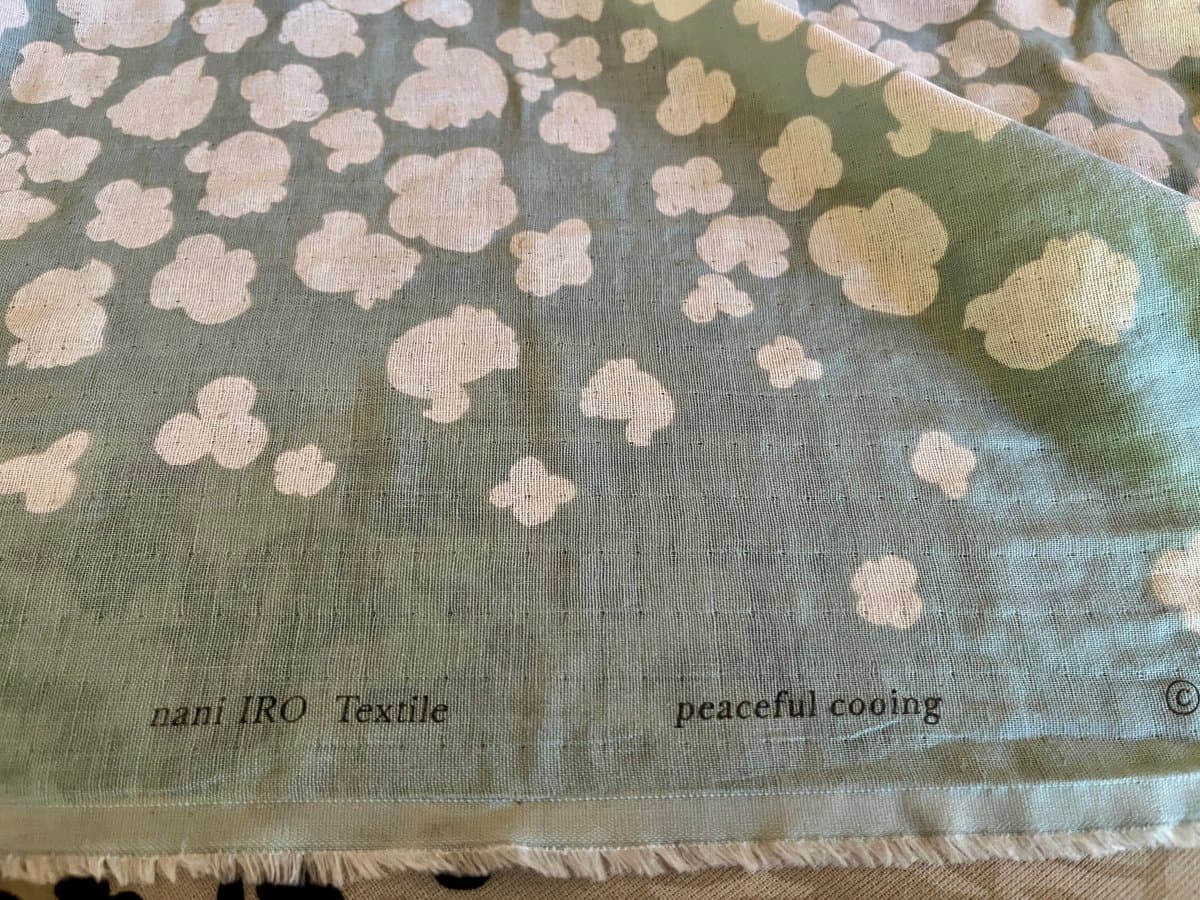 A close-up photograph of a green fabric with a white cloud-like pattern, featuring a hem with printed text and a fringed edge.