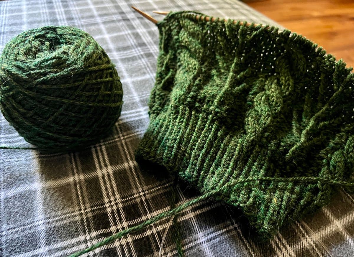 A close-up photograph of a green, textured knitted piece in progress on wooden needles, resting on a plaid tablecloth next to a ball of matching green yarn.