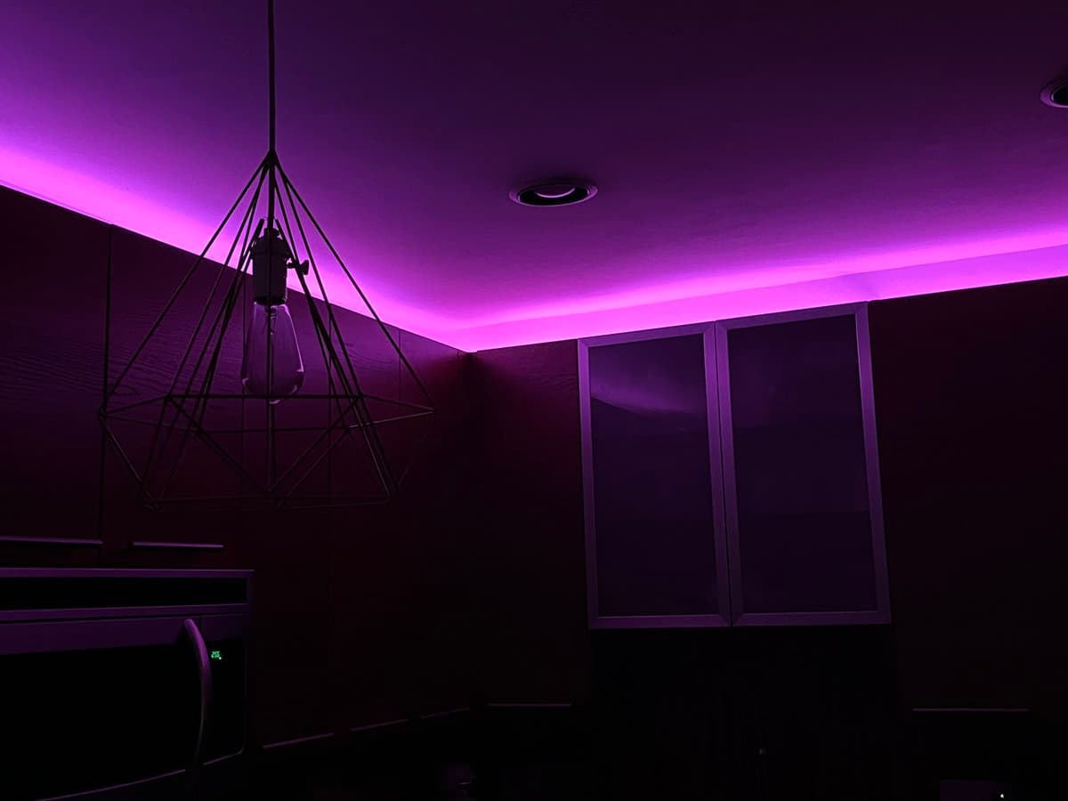 A dimly lit interior room illuminated by purple LED strip lighting along the ceiling cove. A geometric, cage-style pendant light hangs from the ceiling on the left, while a window with closed blinds is visible on the right wall.