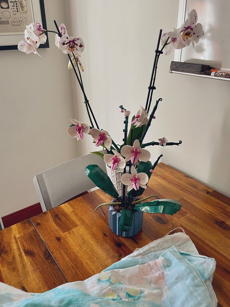 A photograph showing a potted plant on a wooden table that combines real white and pink orchid flowers with a LEGO-built orchid structure in the same pot.