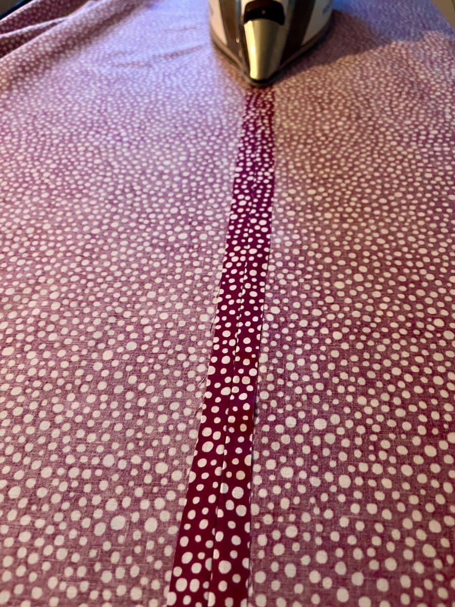 A close-up photograph of a garment with a white polka-dot pattern being ironed, showing a seam and a contrasting darker red polka-dot strip. The tip of a silver and gold iron is visible at the top of the frame.