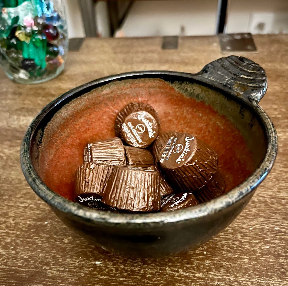 A close-up photograph of a rustic ceramic bowl filled with several wrapped chocolate butter cups sitting on a wooden table. The background is slightly out of focus, showing a glass jar filled with colorful glass beads.