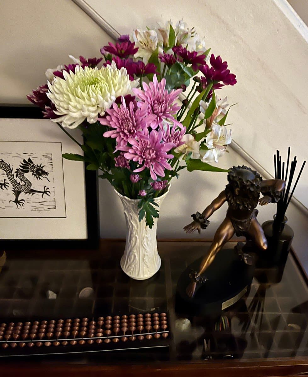 A still-life photograph of a decorative table featuring a white vase filled with pink, purple, and white chrysanthemums, a bronze statue of a figure in motion, a framed black-and-white print of a bird, and a copper Newton's cradle.
